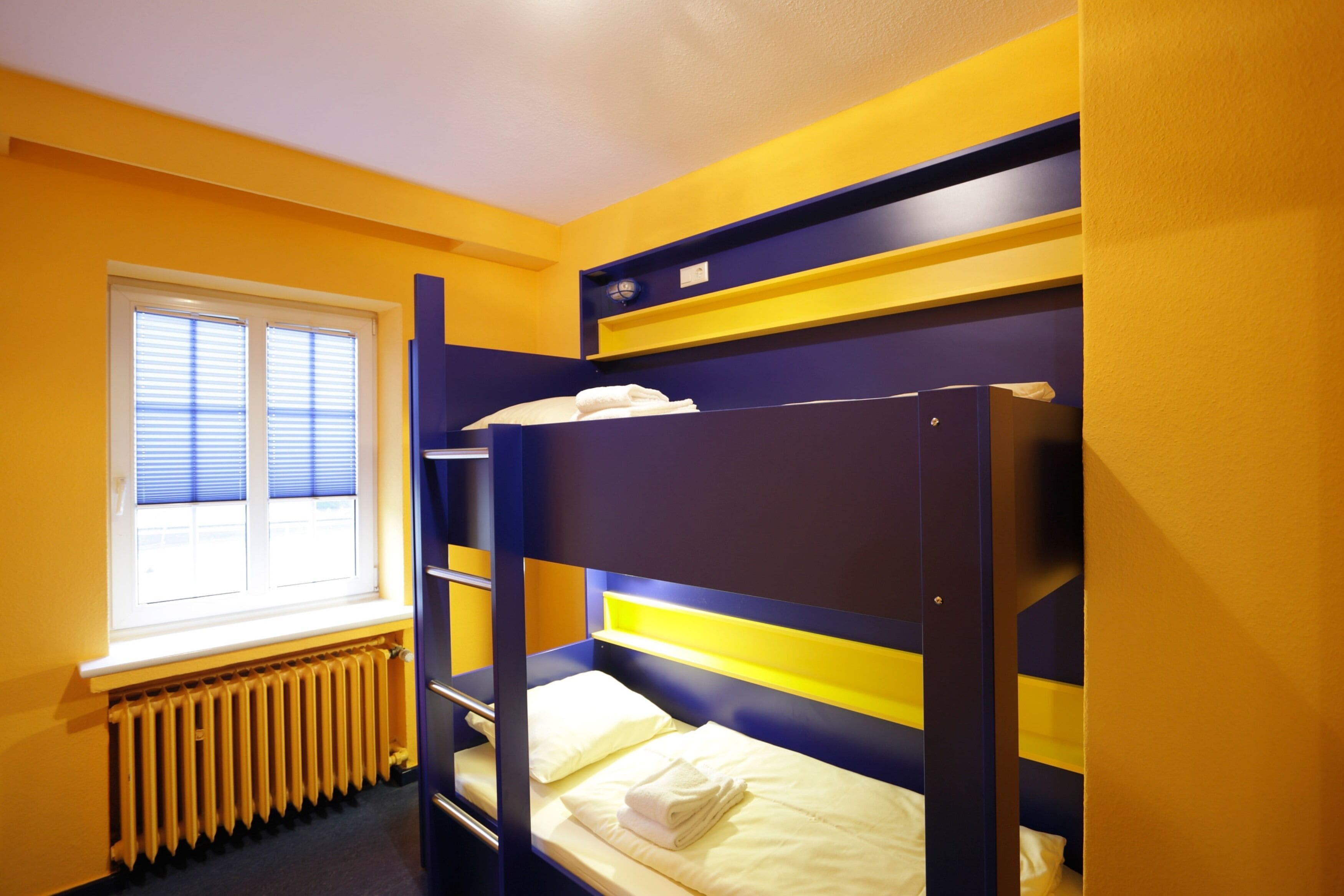 A bunk bed room with yellow walls, a window with blinds, and a radiator.