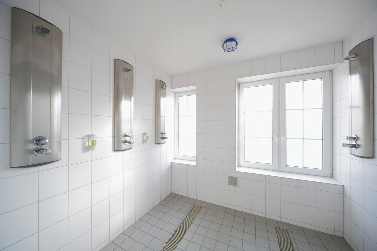 Une salle de bain avec des murs recouverts de carreaux blancs, une fenêtre et une douche dotée d’un panneau de commande.