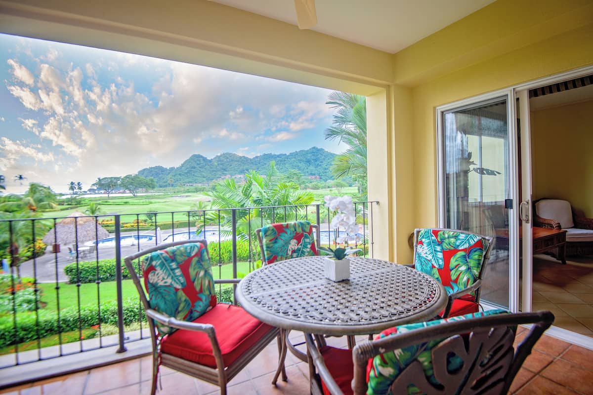 Un balcon avec une table et des chaises, une vue sur un paysage verdoyant et une porte donnant sur un espace intérieur.