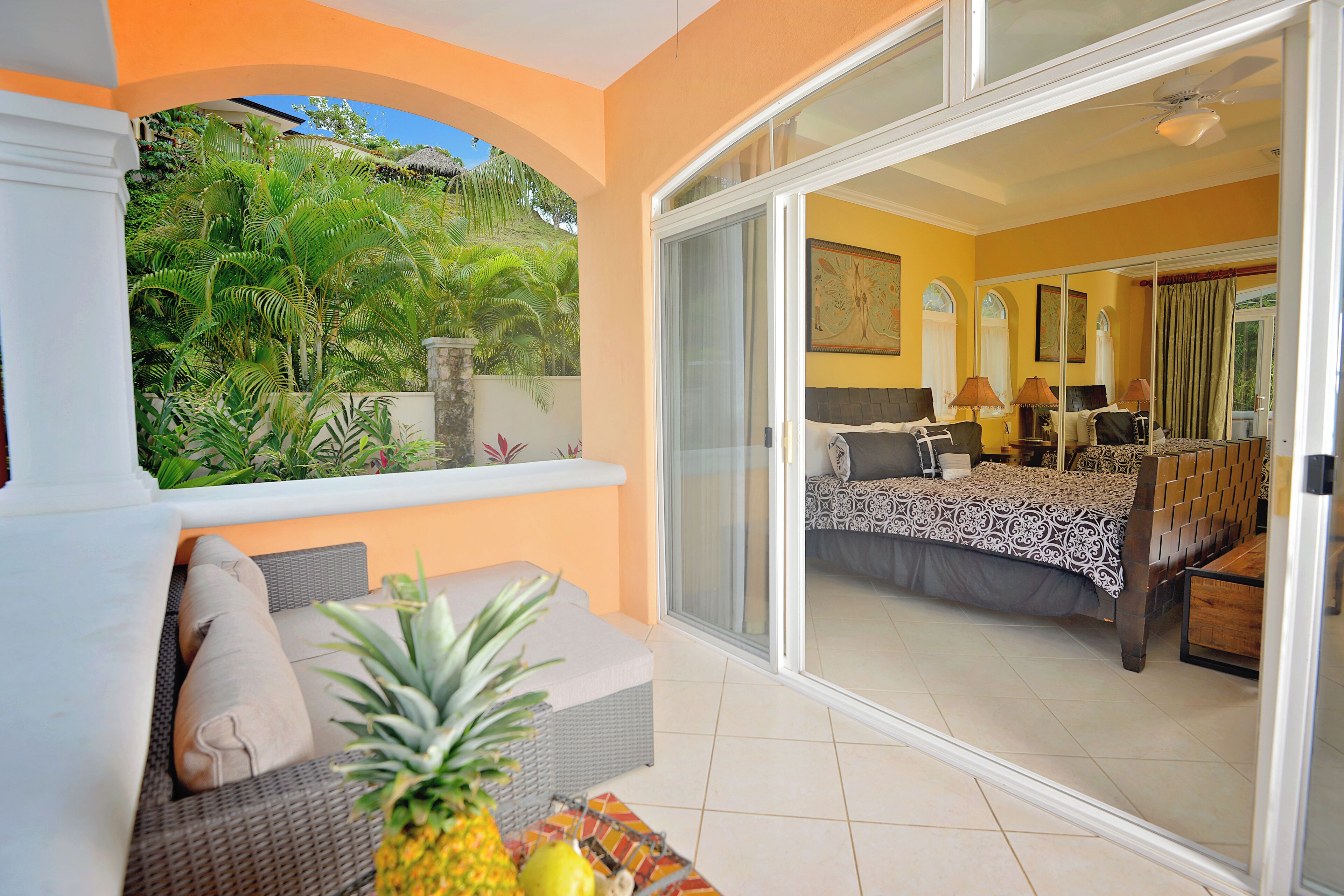 A balcony with a wicker sofa, a dining table with a pineapple, and a view of a bedroom with a bed and a window.