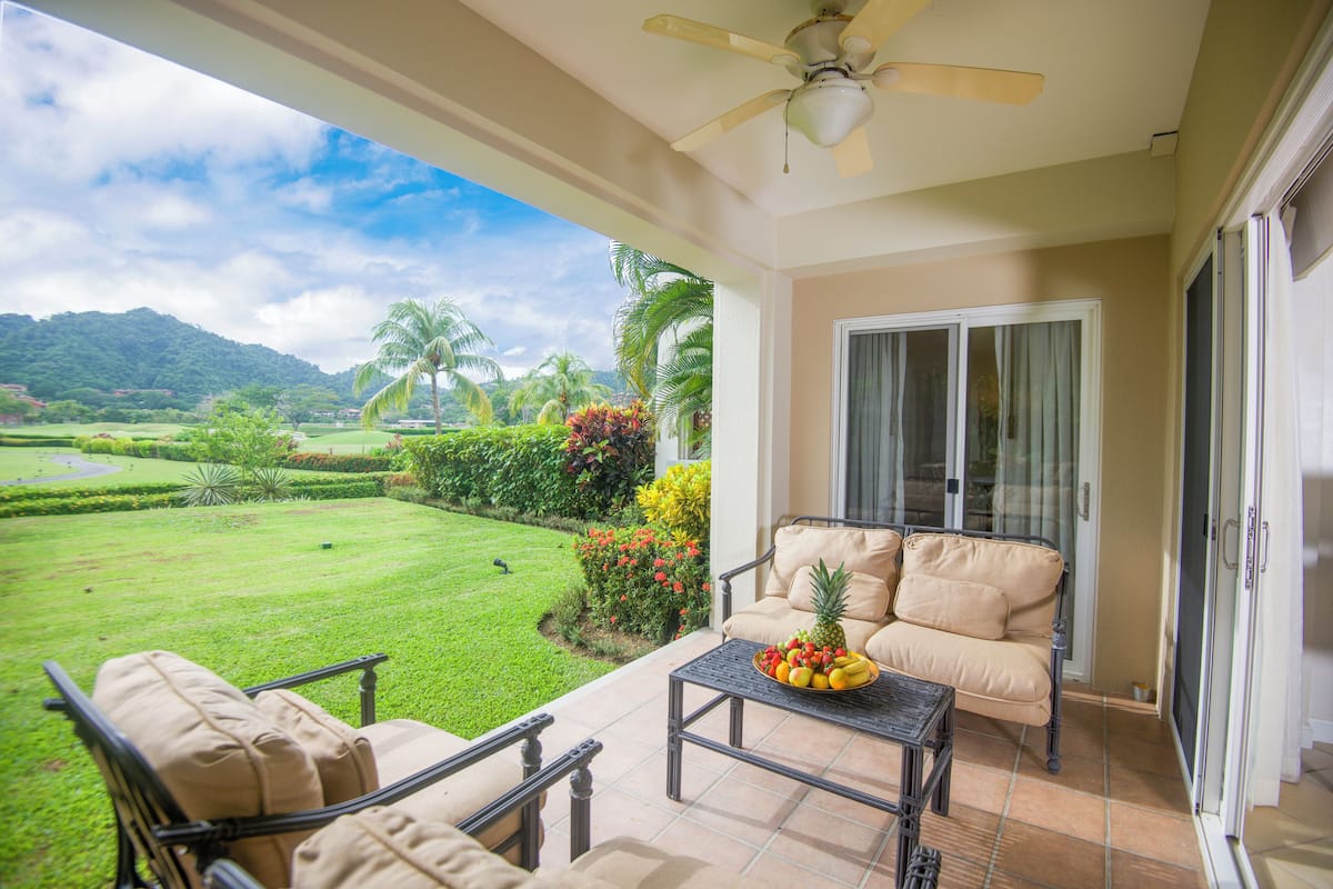 Un balcon donnant sur un jardin luxuriant et les montagnes, un ventilateur de plafond et une table avec des fruits.