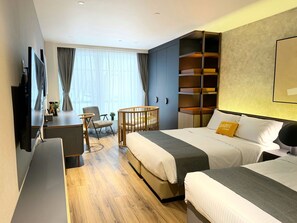 Une chambre d’hôtel avec deux lits, un bureau, une chaise, un berceau et une grande fenêtre avec des rideaux.