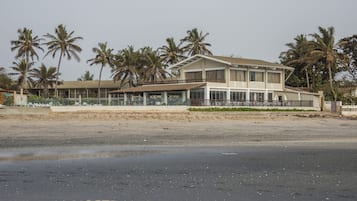 Ein Strandhotel mit einem großzügigen, zweistöckigen Gebäude, einer überdachten Terrasse und Palmen im Hintergrund.