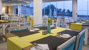 A restaurant with white chairs and tables, yellow tablecloths, and a view of palm trees through large windows.
