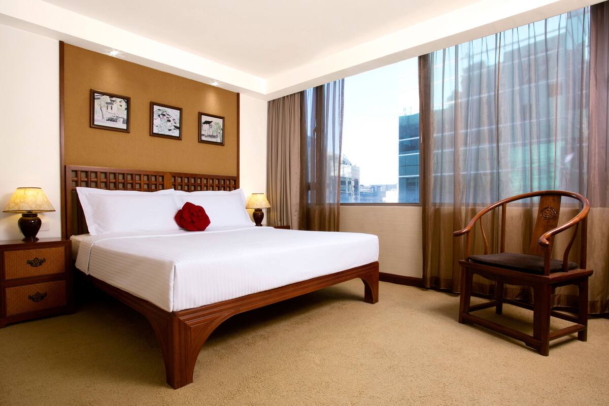 A hotel room with a large bed, two bedside tables with lamps, a wooden chair, and a view of the city through large windows.