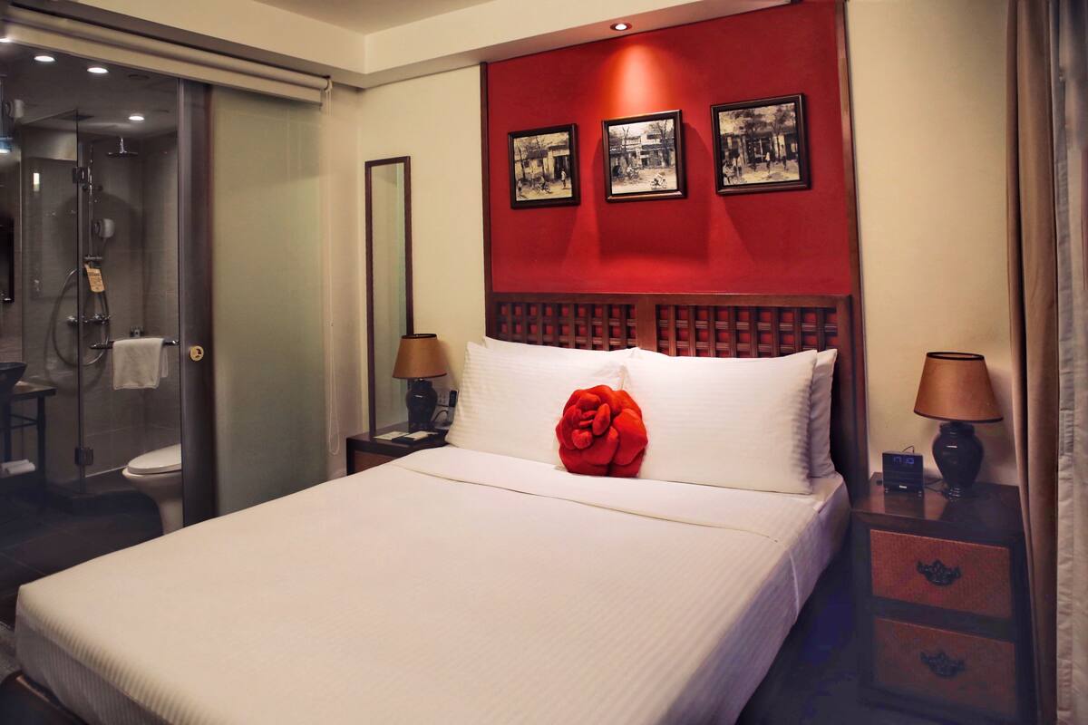 A hotel room with a large bed, a wooden headboard, a glass shower enclosure, and a nightstand with a lamp.