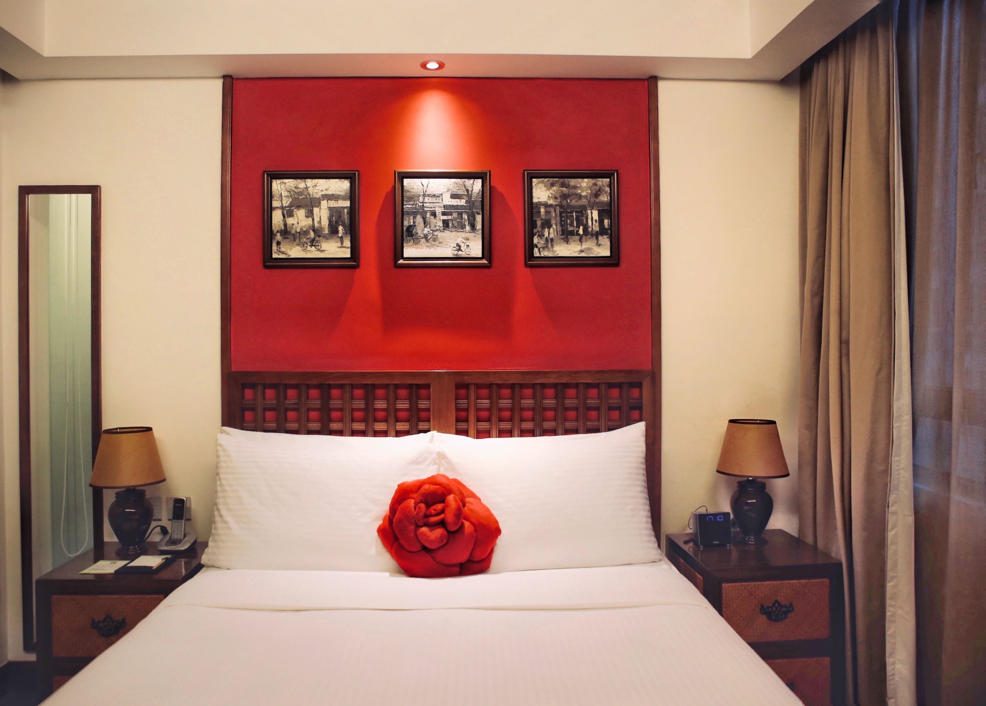 A hotel room with a bed, a red accent wall, three framed pictures, two bedside lamps, and a red rose decoration.