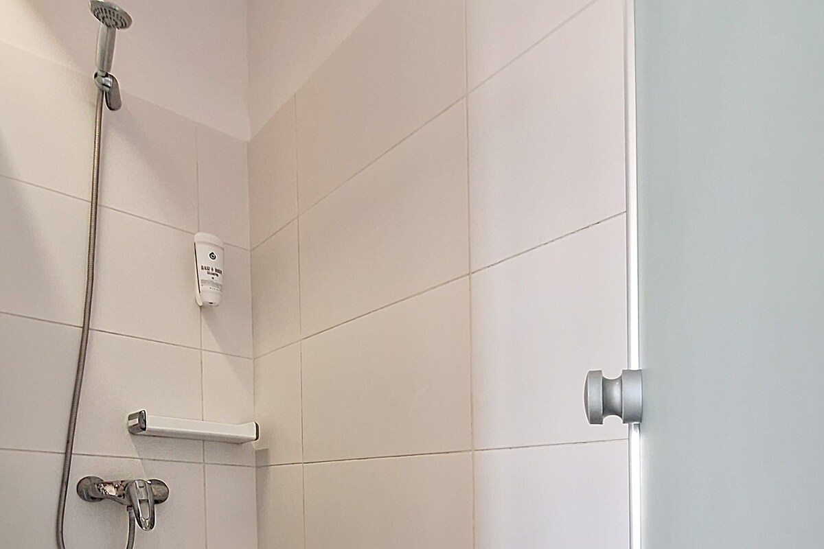 A bathroom with white tiled walls, a shower with a handheld showerhead, a towel rack, and a door with a round handle.
