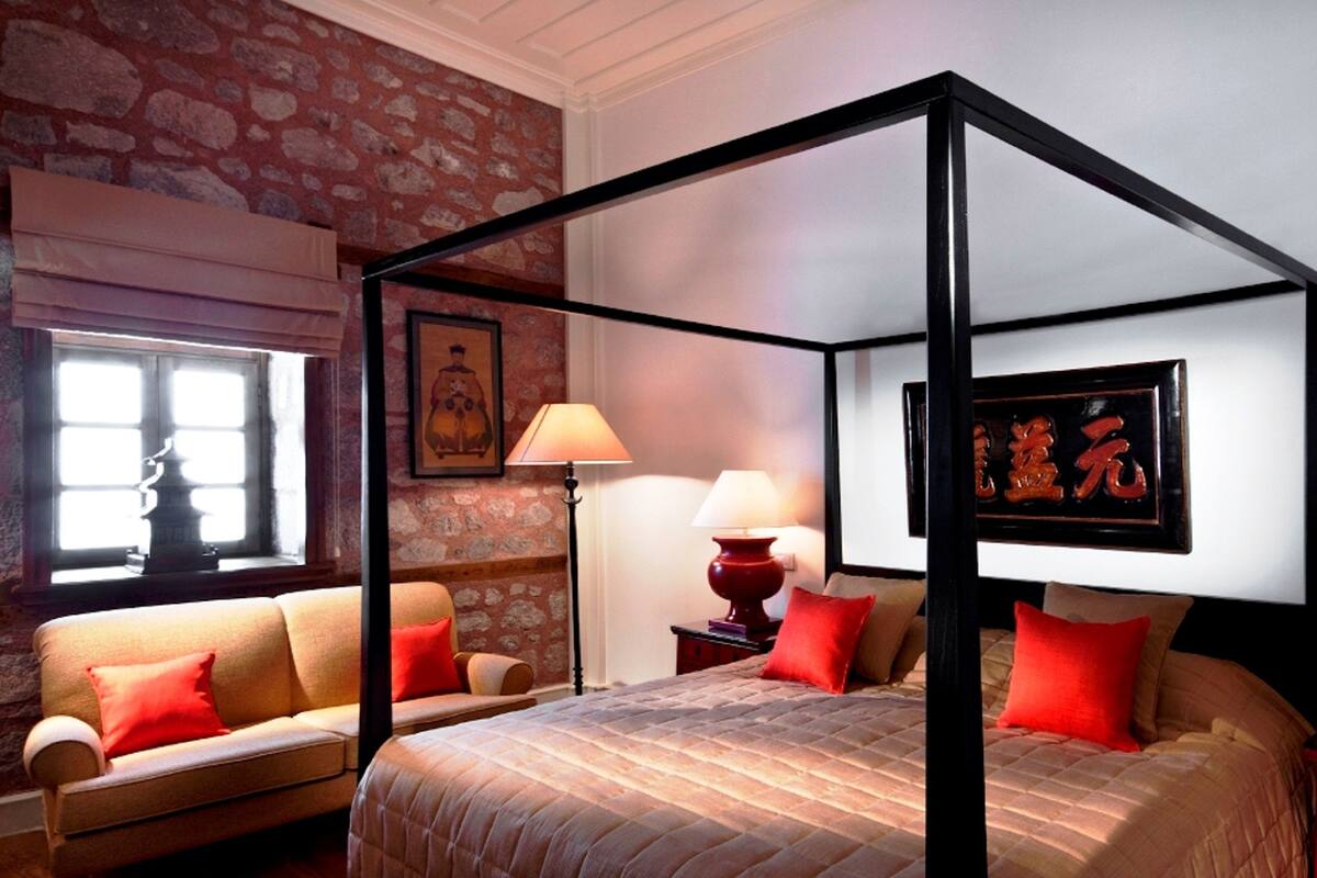 Ein Bett mit Baldachin, rote Kissen, eine Couch mit roten Kissen und ein Fenster mit einer dekorativen Lampe.