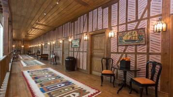 A hallway with wooden floors, patterned walls, and a long rug. There are chairs, a table, and wall-mounted lights.