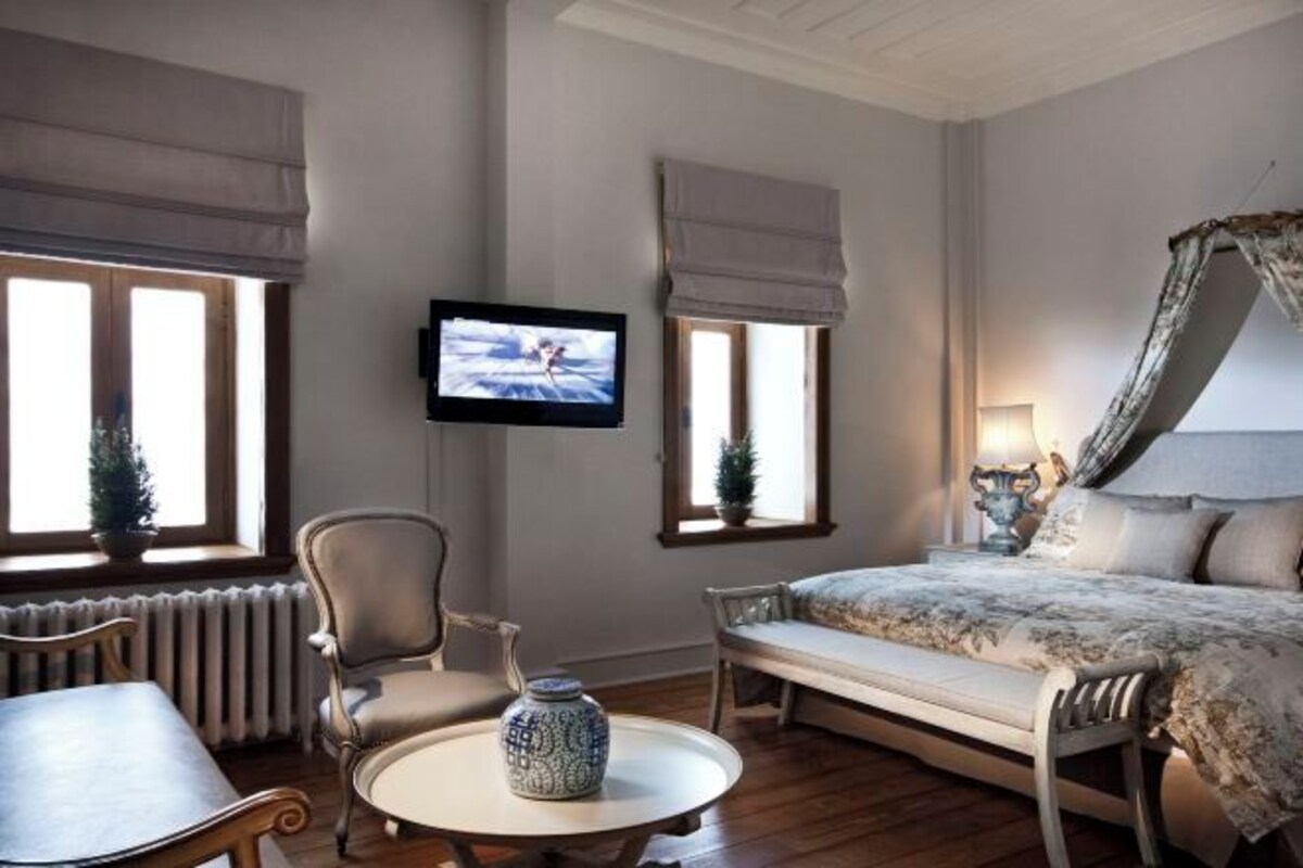 A bedroom with a bed, a TV, a chair, a table, and a radiator.