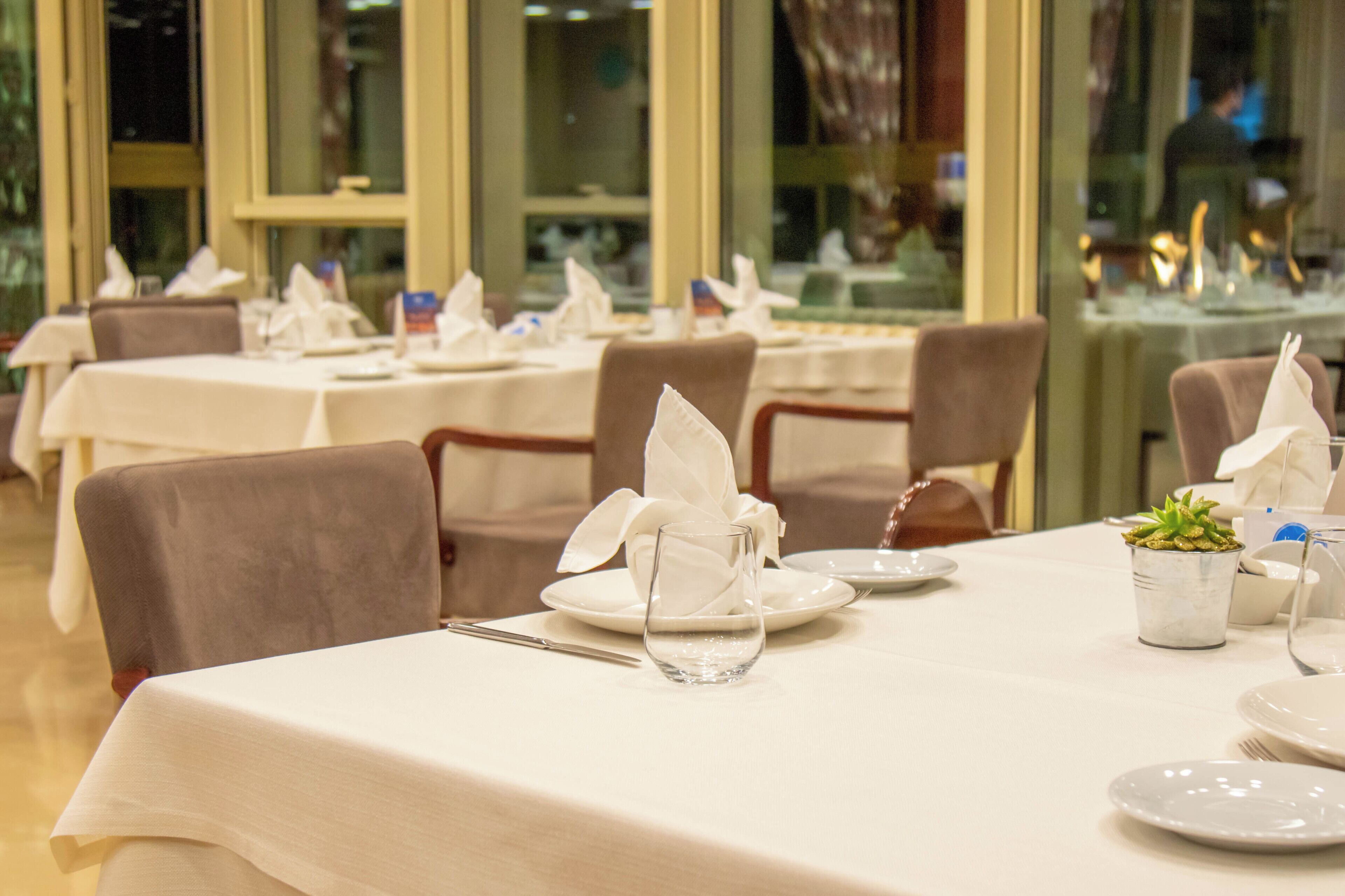 A restaurant with tables set for dining, featuring white tablecloths and neatly arranged plates, glasses, and napkins.