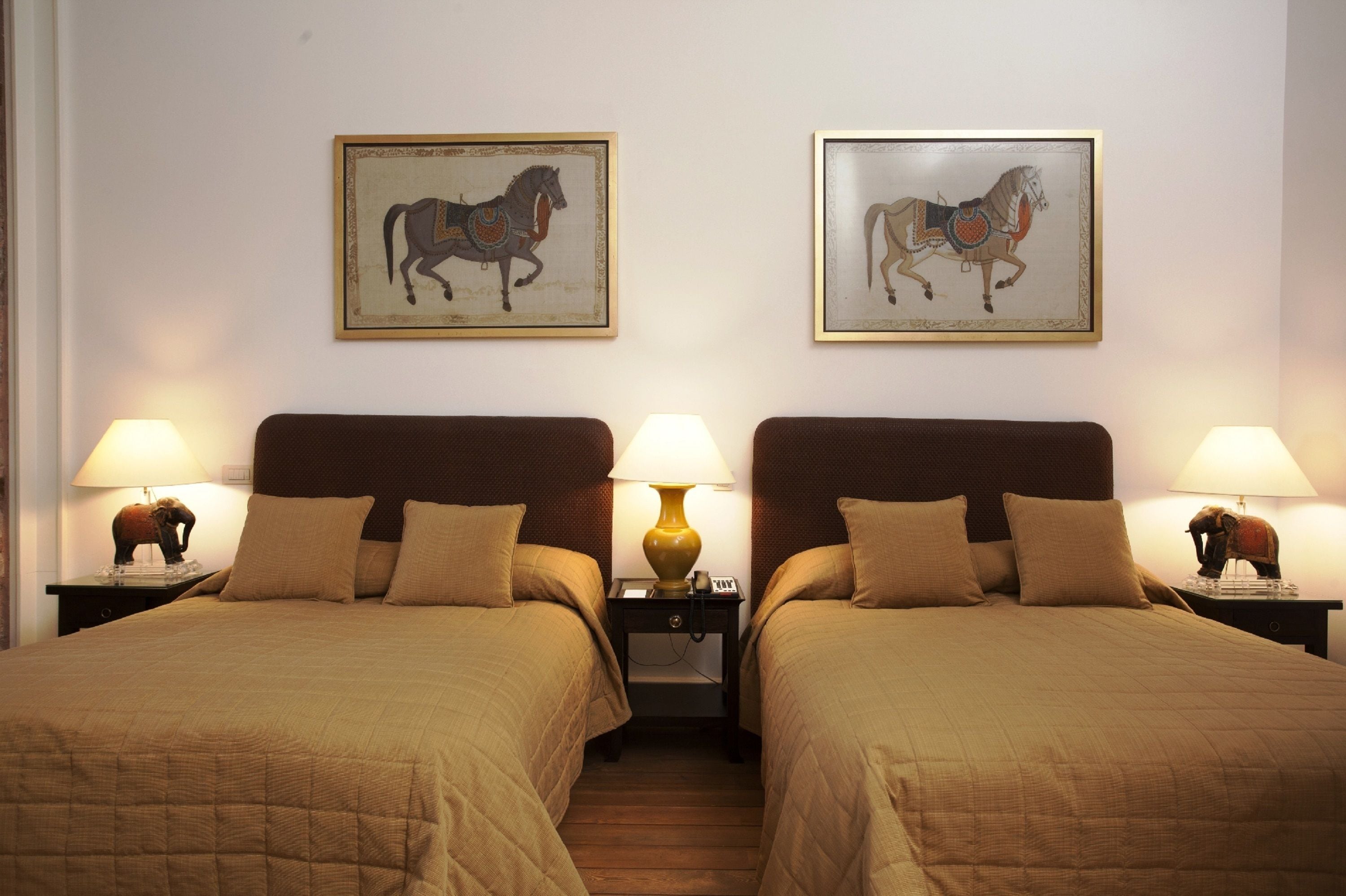 A hotel room with two beds, each with a headboard, and two lamps on nightstands. There are two framed pictures of horses on the wall.