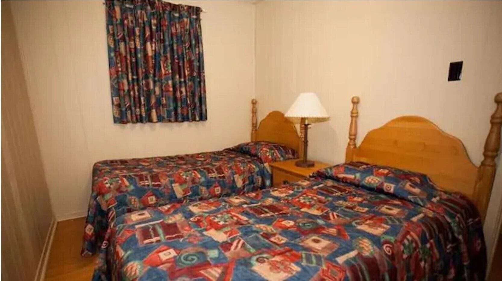 A room with two single beds, each with a colorful quilt, a bedside table with a lamp, and a curtain with a pattern.