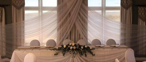 A banquet hall with a long table set for a formal event, draped with white fabric and a floral centerpiece.