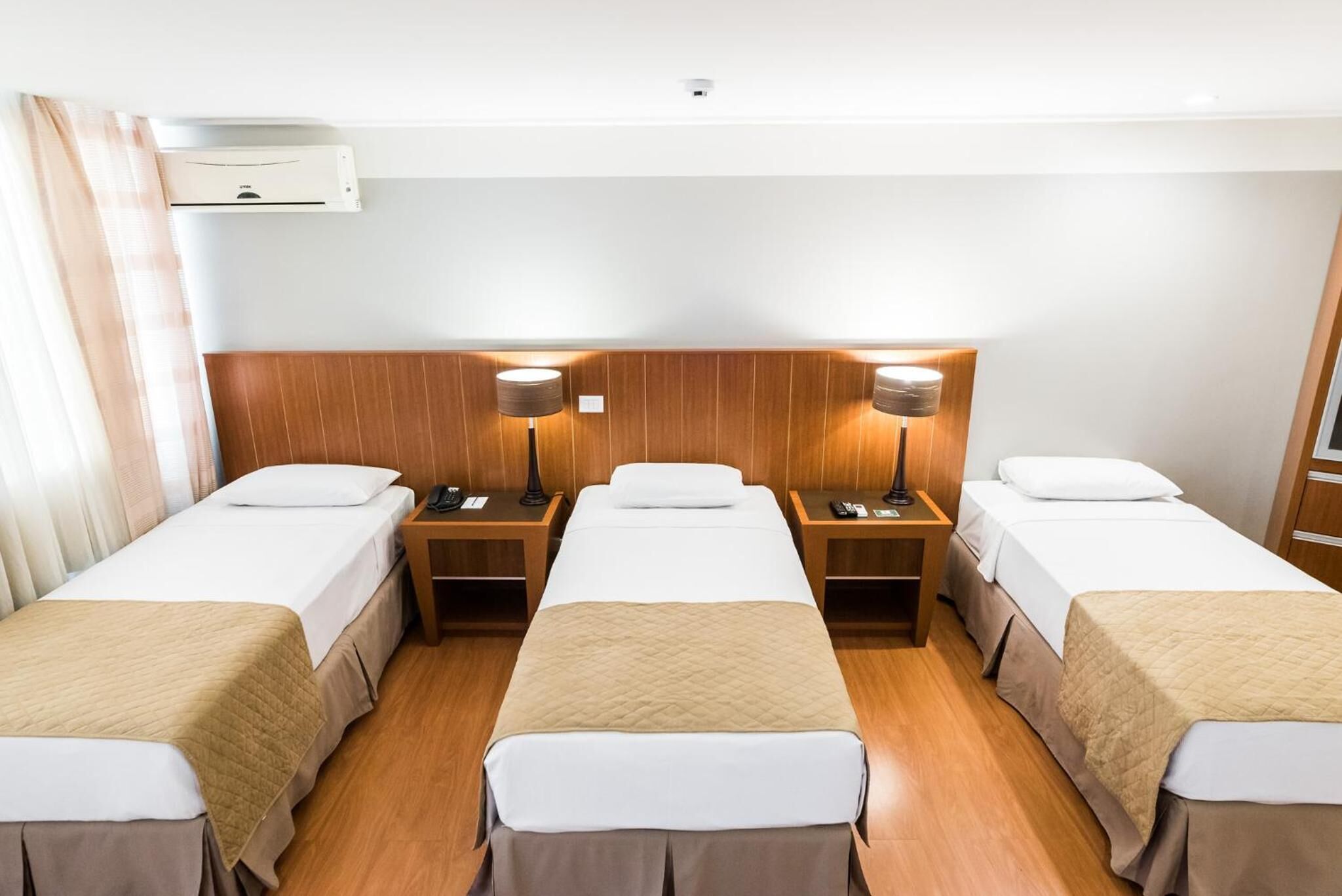 A hotel room with four single beds, wooden headboards, bedside tables with lamps, and an air conditioning unit on the wall.
