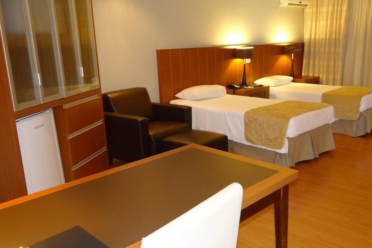 A hotel room with two beds, a desk, a chair, a small refrigerator, and a wall-mounted lamp.