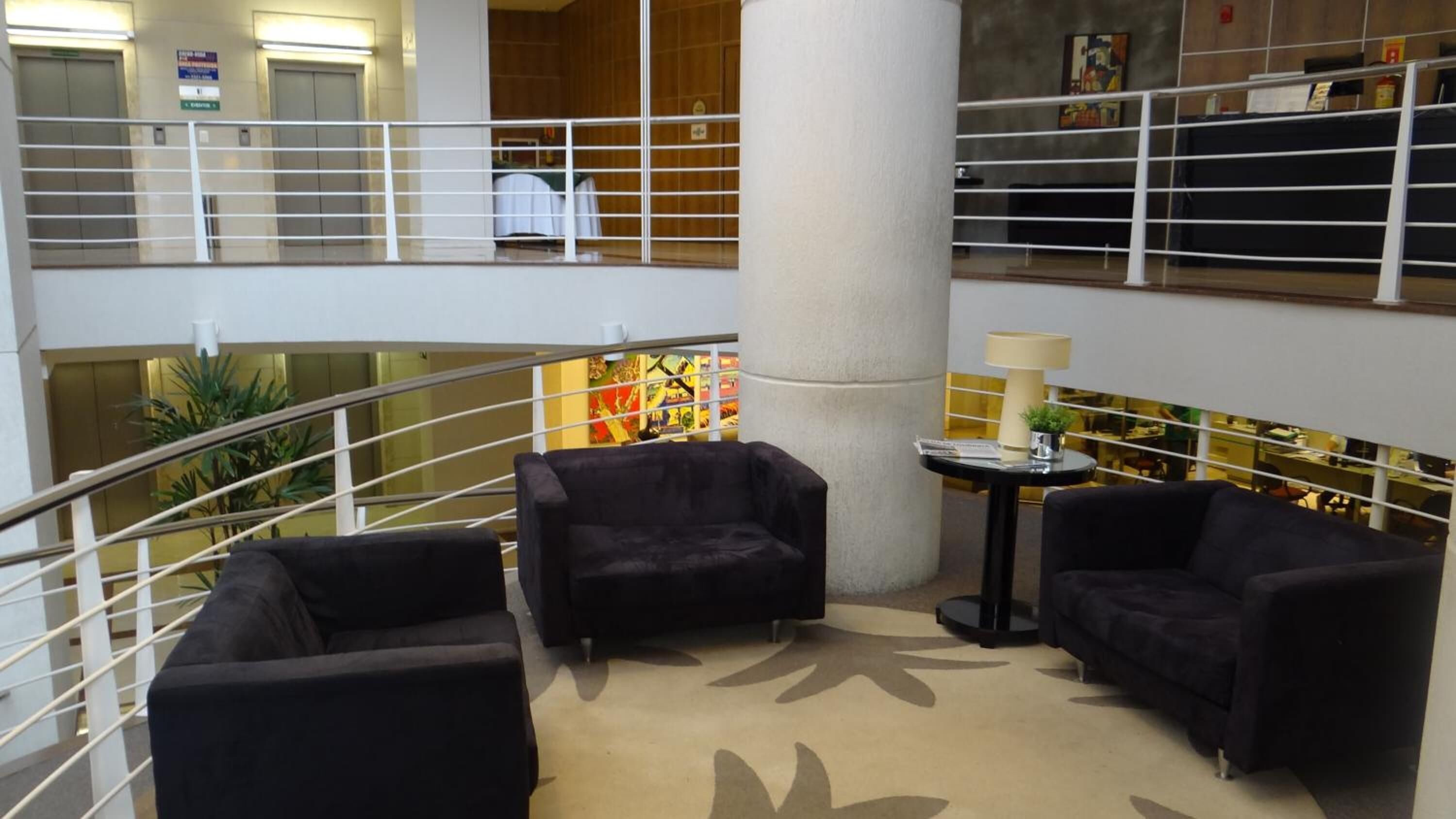 A hotel lobby with a central column, two black armchairs, a small table with a lamp, and a staircase with a railing.