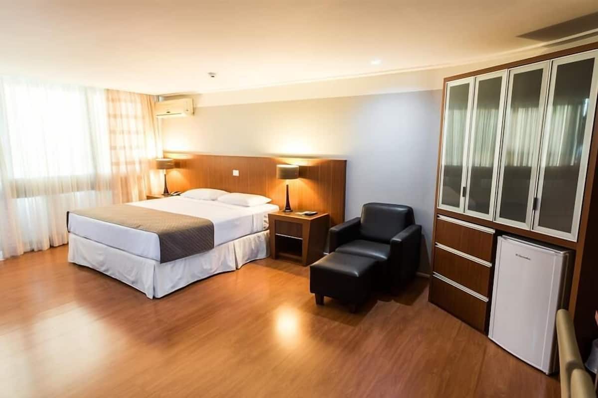 A hotel room with a double bed, a small desk, a chair, a wardrobe, and a mini-fridge.