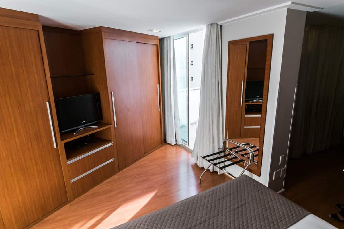 A modern hotel room with a wooden wardrobe, a flat-screen TV, a bed, and a window with curtains.