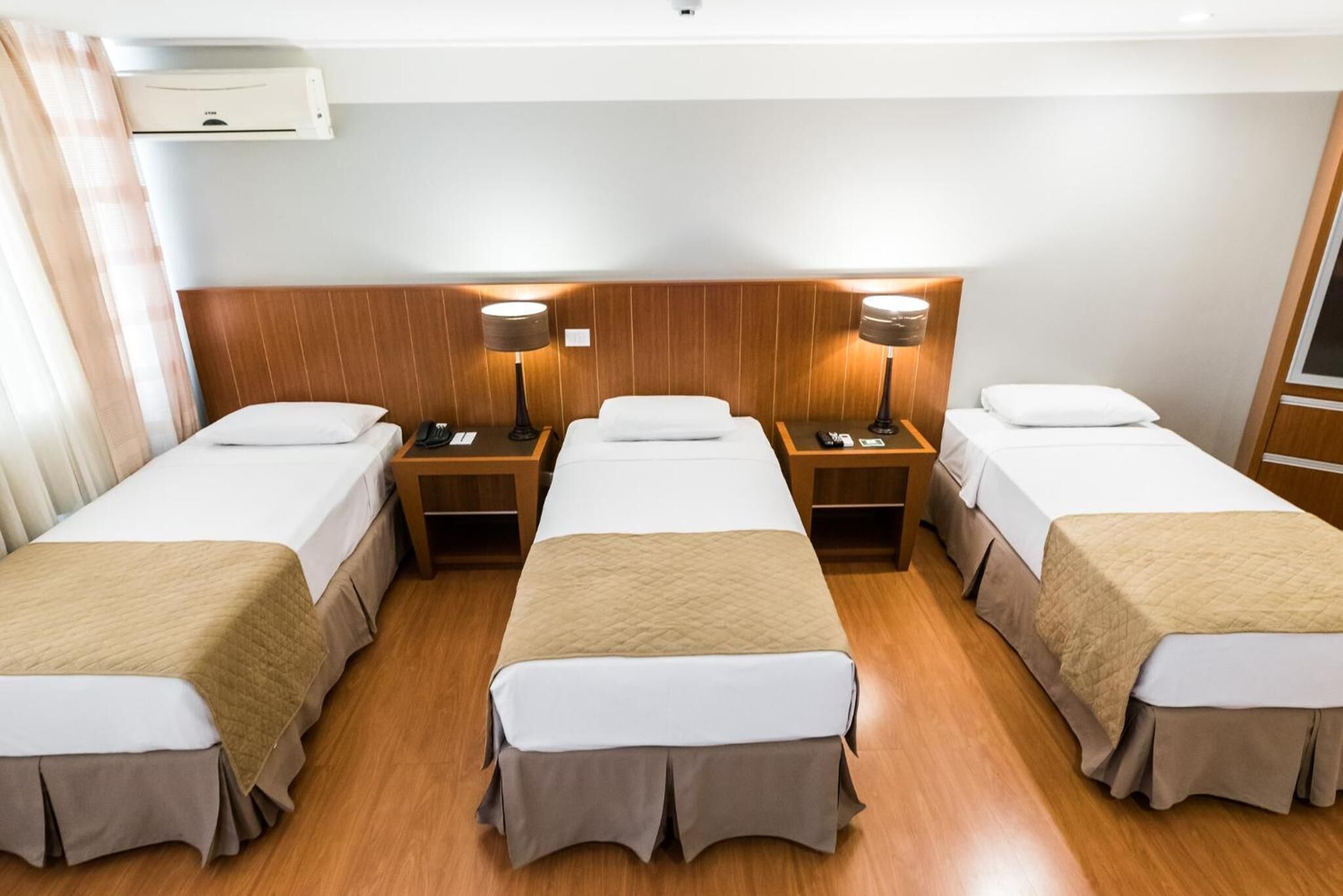 Habitación de hotel con tres camas individuales, cabeceros de madera, mesitas de noche con lámparas y un equipo de aire acondicionado en la pared.