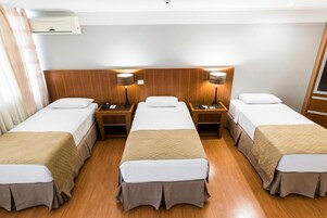 Habitación de hotel con tres camas individuales, cabeceros de madera, mesitas de noche con lámparas y un equipo de aire acondicionado en la pared.