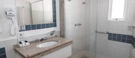 A bathroom with a granite countertop sink, a mirror, a hairdryer, and a glass-enclosed shower.