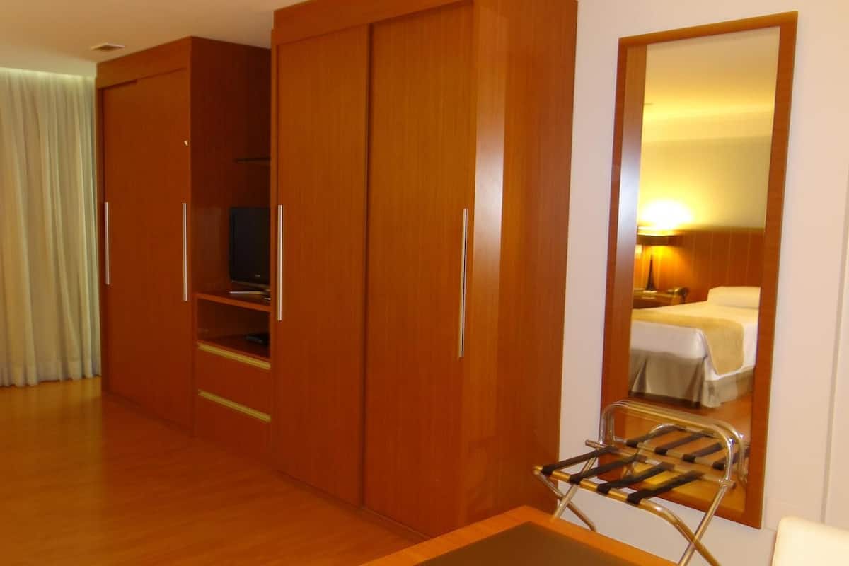 A hotel room with a wooden wardrobe, a television, a mirror reflecting a bed, and a small table with a chair.