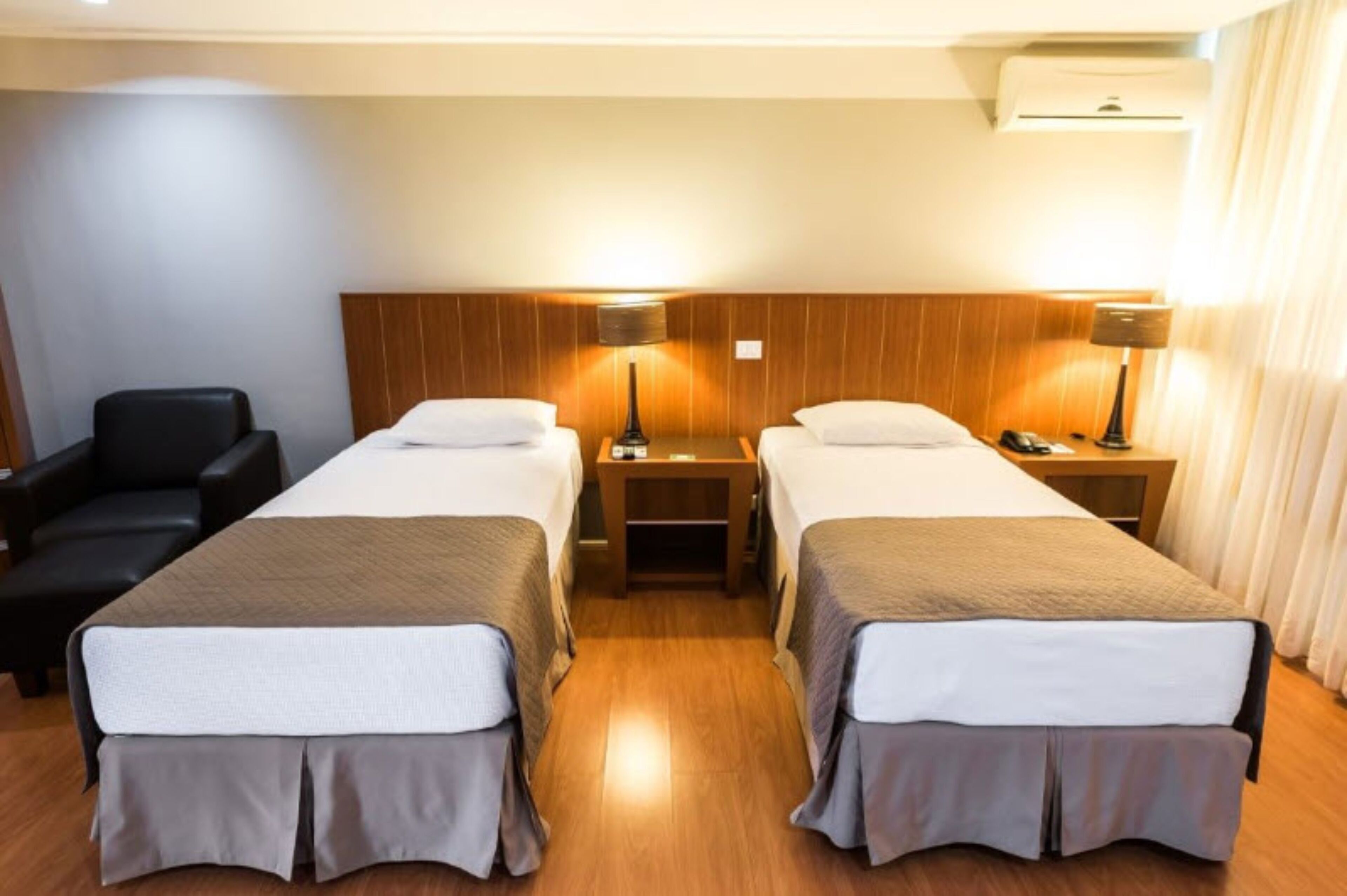 A hotel room with two single beds, a wooden headboard, bedside tables with lamps, a black leather chair, and an air conditioning unit.