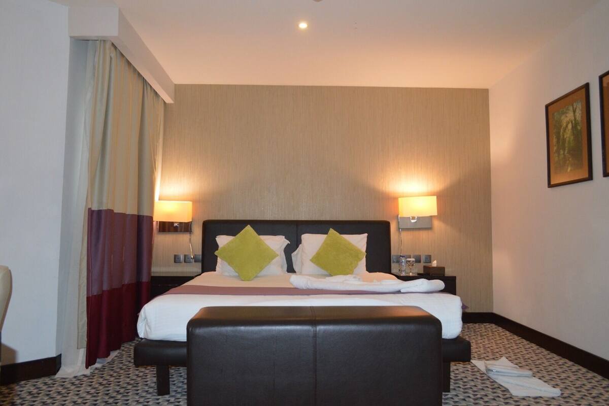Quarto de hotel com cama, duas luminárias de cabeceira, mesa de cabeceira, uma cadeira e carpete estampado.