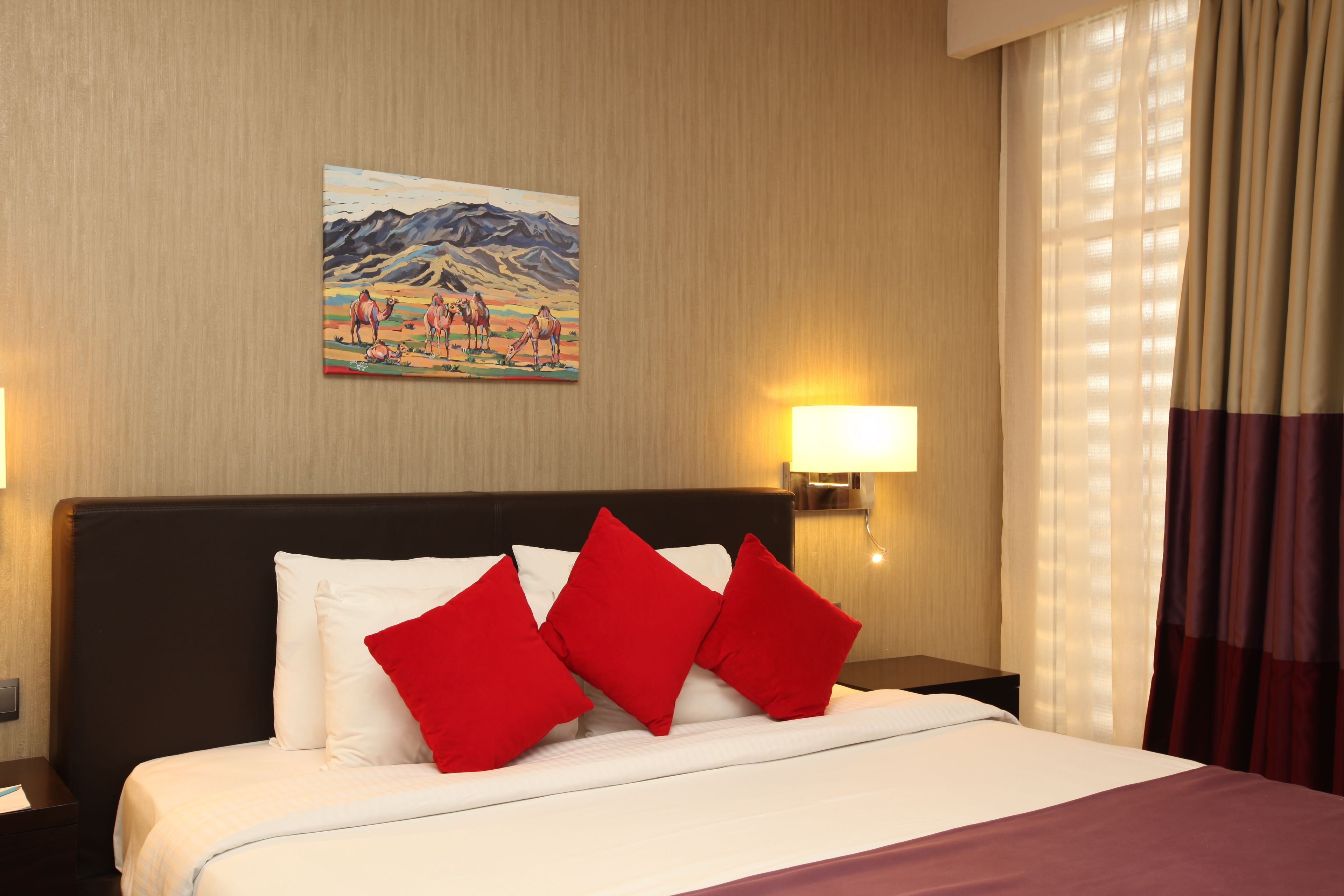A hotel room with a large bed, red pillows, a painting on the wall, and two bedside lamps.