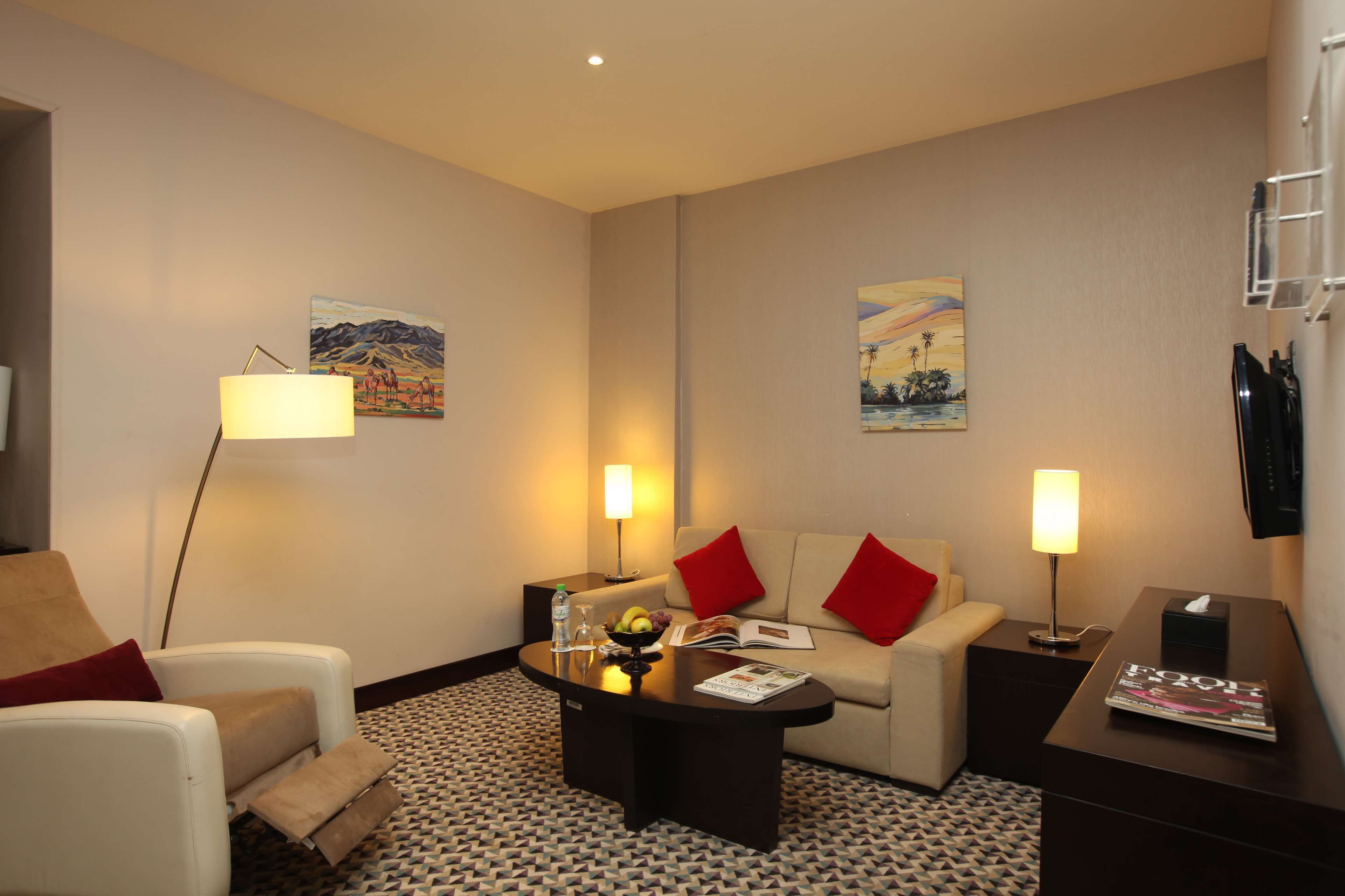 Een hotelkamer met een bank, fauteuil, salontafel en twee nachtkastjes met lampen. Op de muren hangen twee schilderijen en er ligt een tijdschrift op de salontafel.