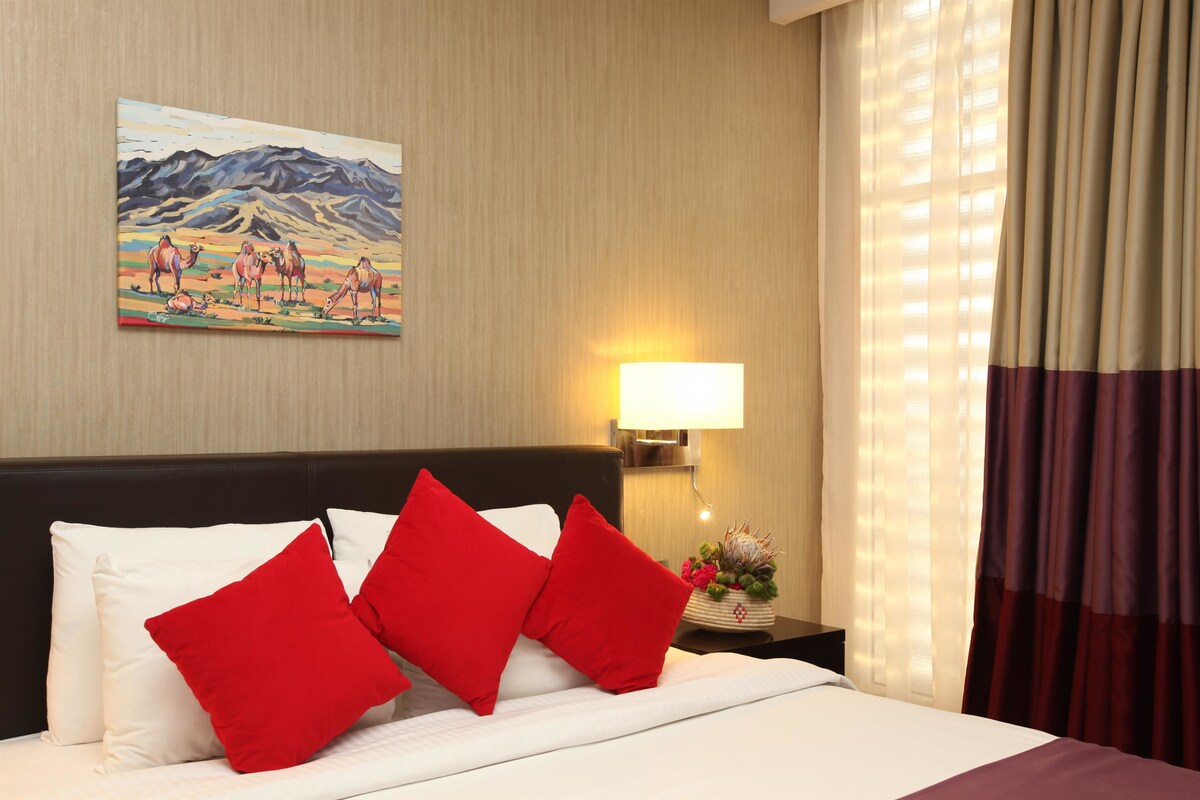 A hotel room with a bed, red pillows, a nightstand with a lamp, a painting on the wall, and curtains.