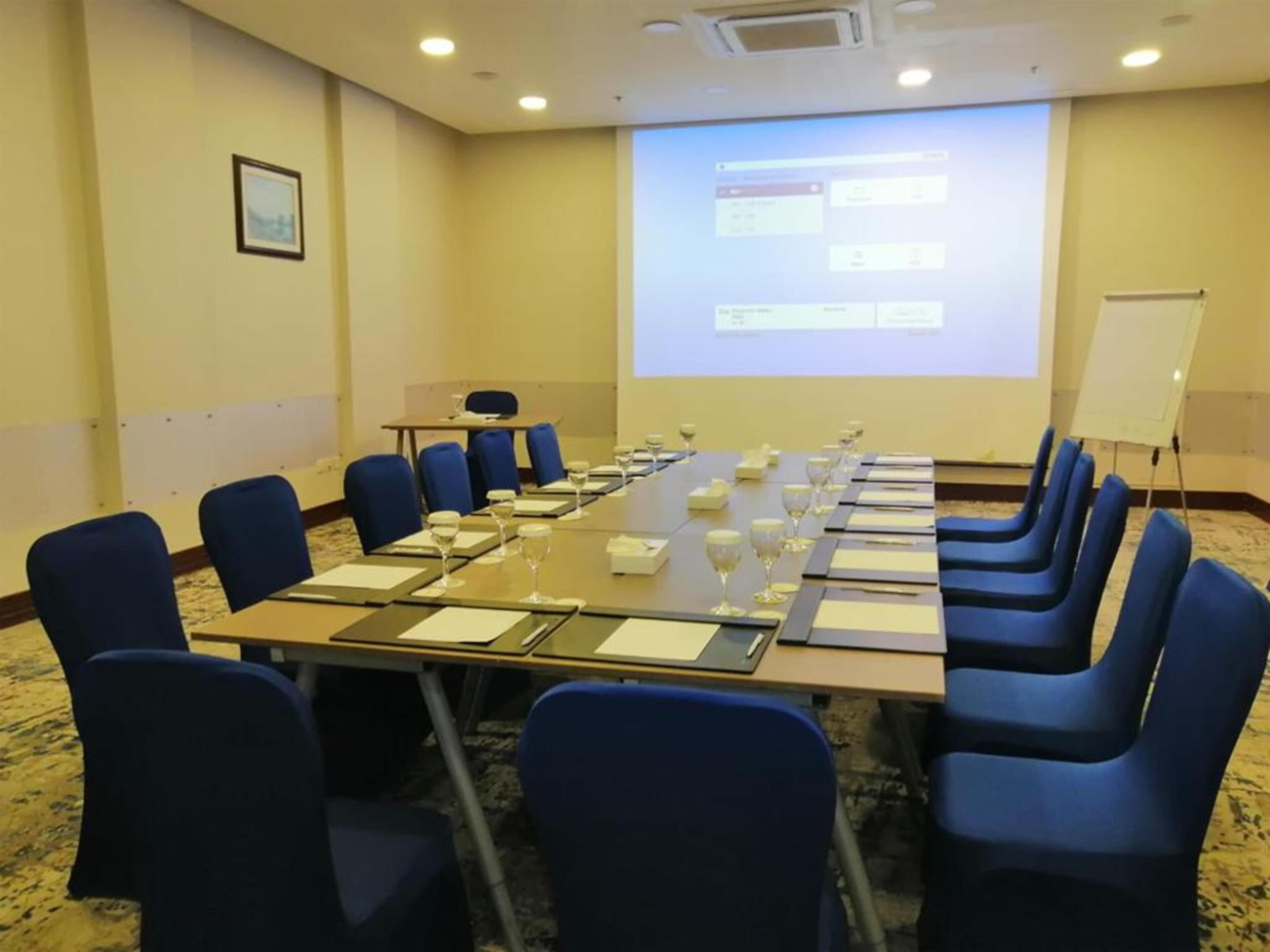 Un salón de conferencias con una larga mesa, sillas azules y una pantalla que muestra una presentación.