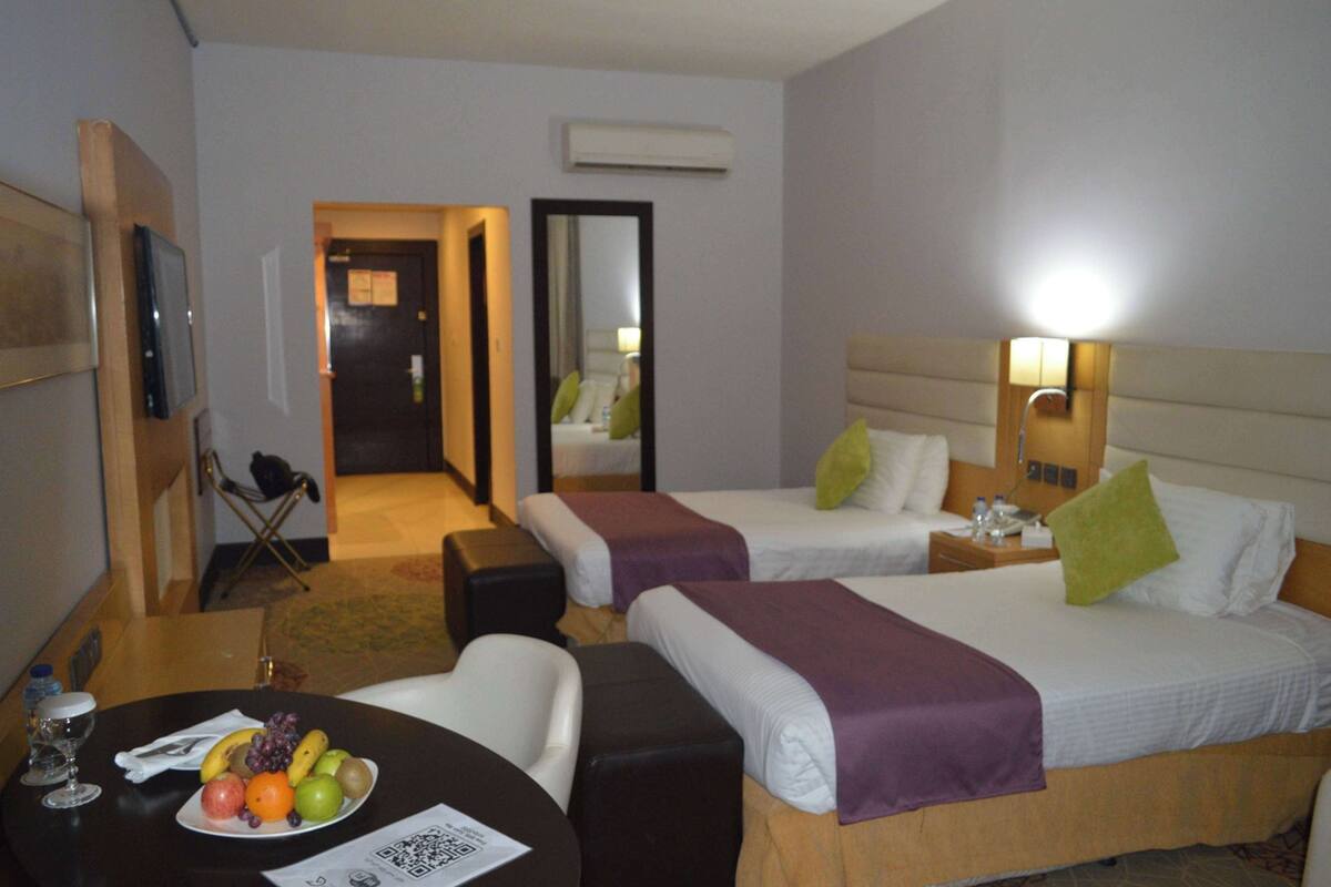 Quarto de hotel com duas camas, uma mesa com uma cesta de frutas, uma cadeira e uma televisão.