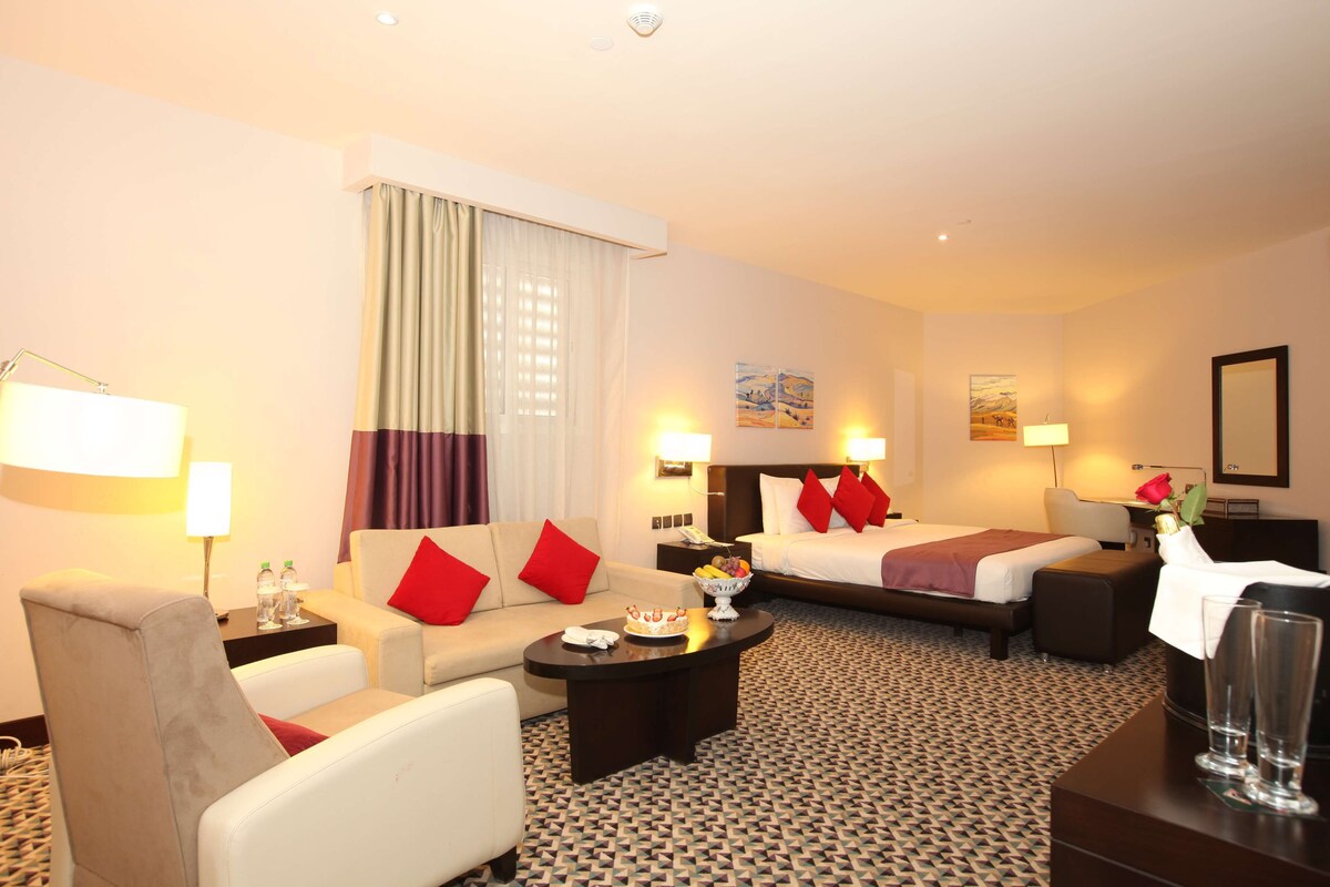 A hotel room with a sofa, a bed, a coffee table, and a desk. There are two lamps, a mirror, and a painting on the wall.