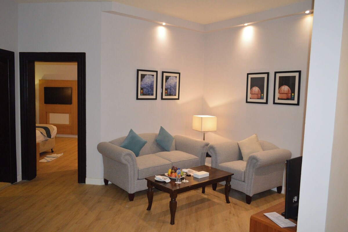 A living room with two sofas, a coffee table, and a TV. There are framed pictures on the wall and a lamp on the table.