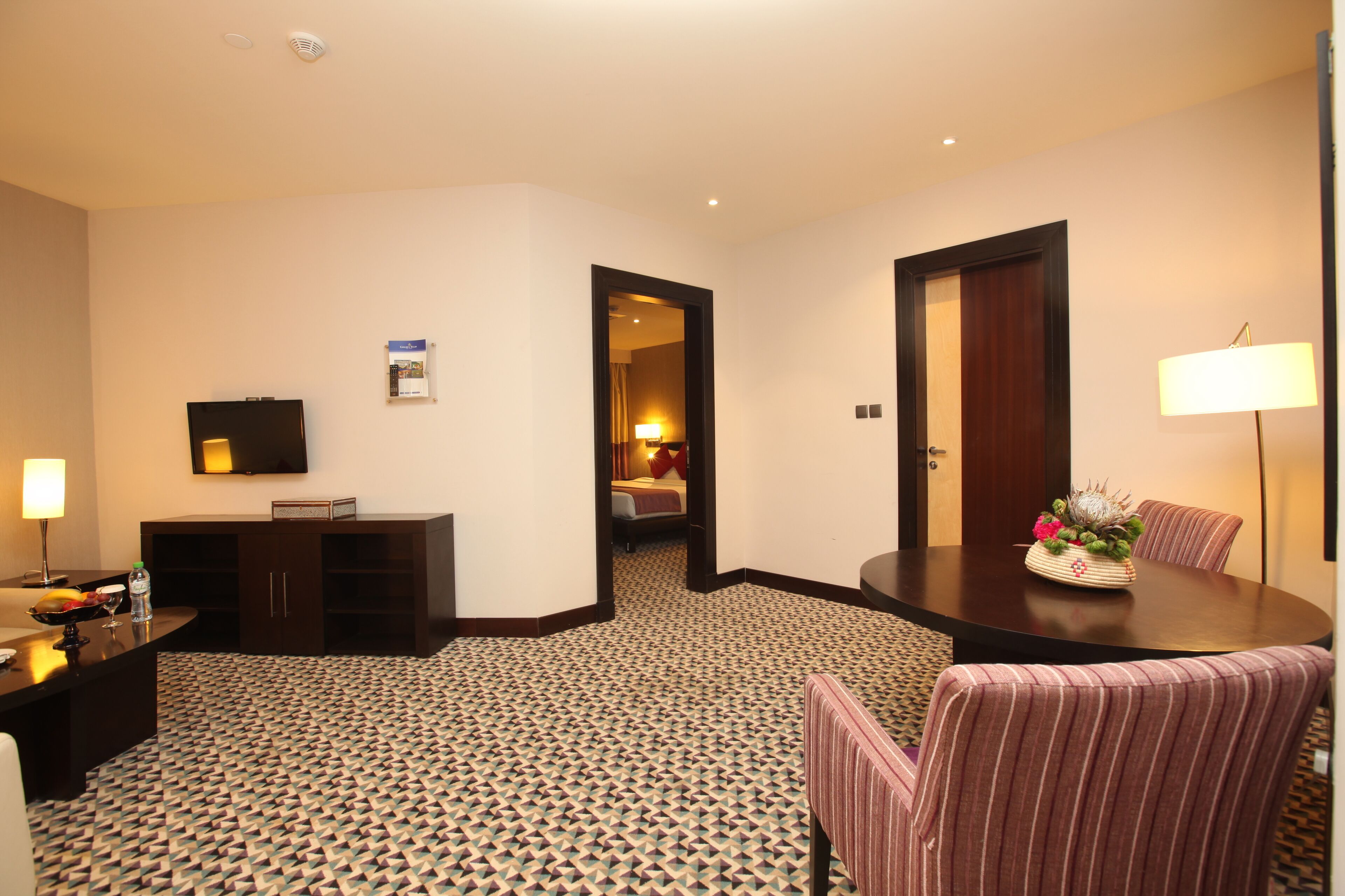 Habitación de hotel con zona de comedor, televisor, minibar y alfombra estampada.