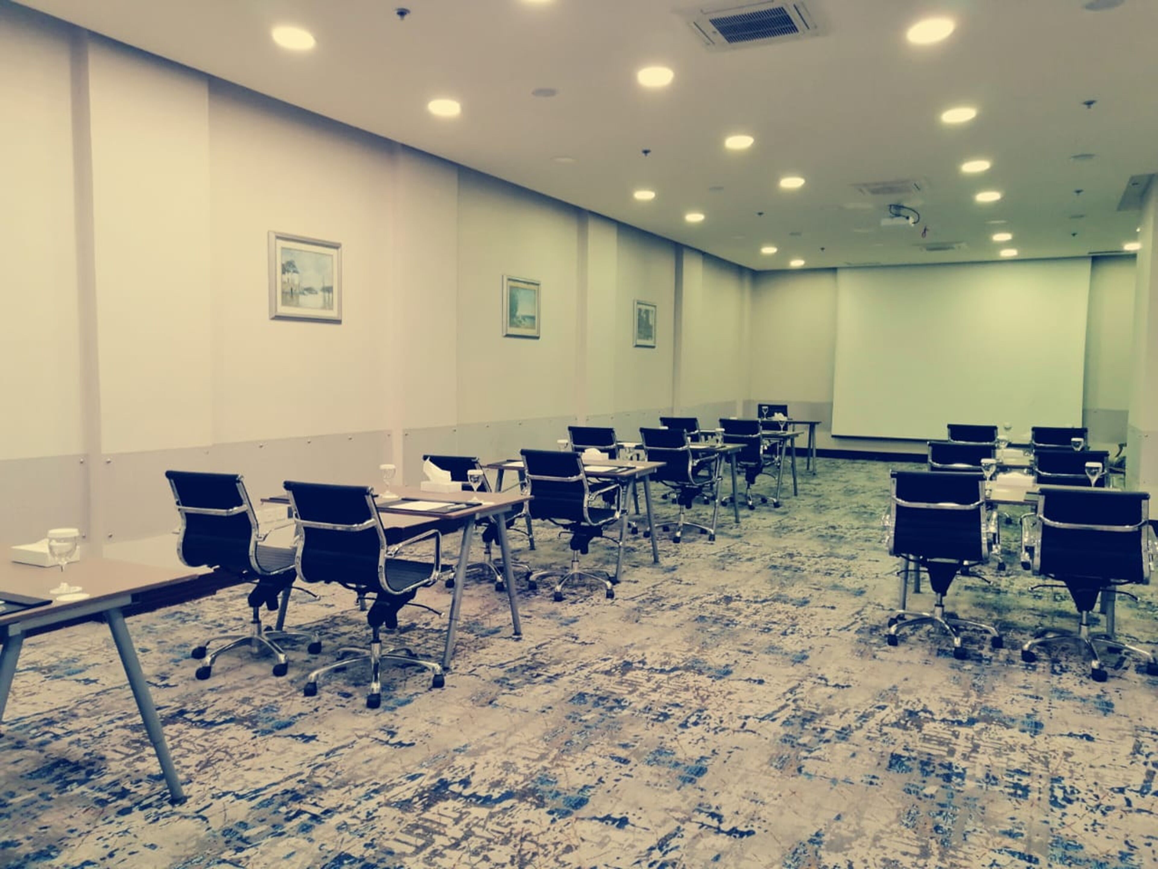 Una sala de conferencias con varias mesas y sillas, piso alfombrado y una pared lisa con pantalla de proyector.