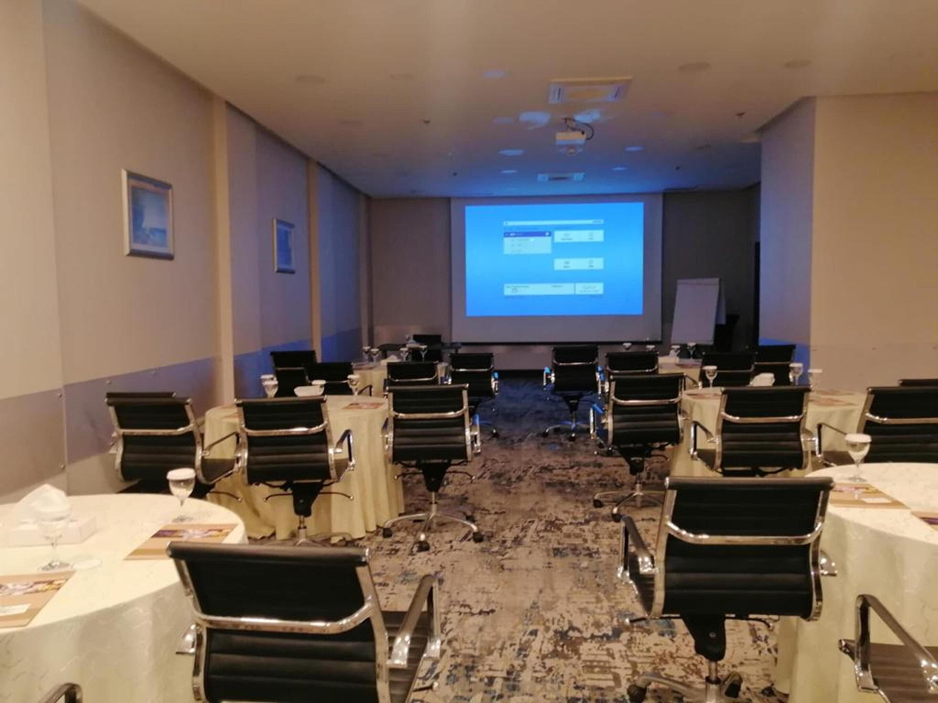 Ein Konferenzraum mit mehreren Tischen und Stühlen, einer großen Leinwand im Vordergrund und einem Teppichboden.