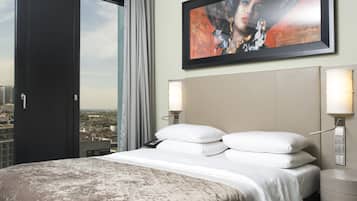A hotel room with a large bed, a city view through a window, and a framed artwork on the wall.