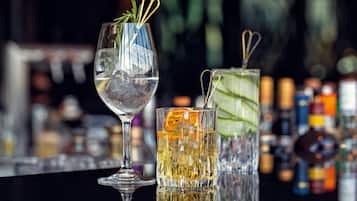 Two glasses of beverages, one with ice and the other with a slice of orange, on a bar counter with a blurred background of bottles.