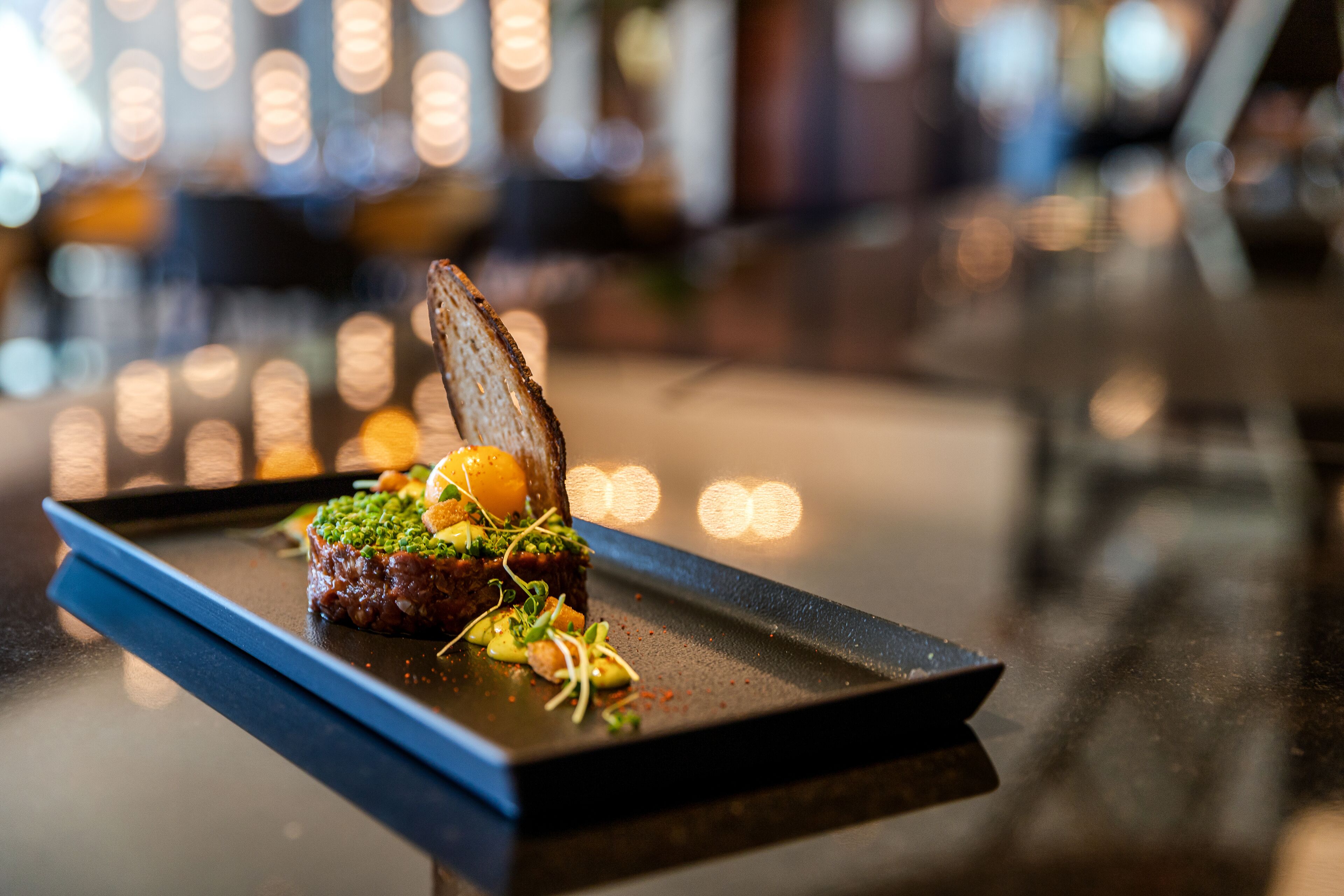 A plated dish with a piece of meat, greens, and a slice of bread on a rectangular plate.