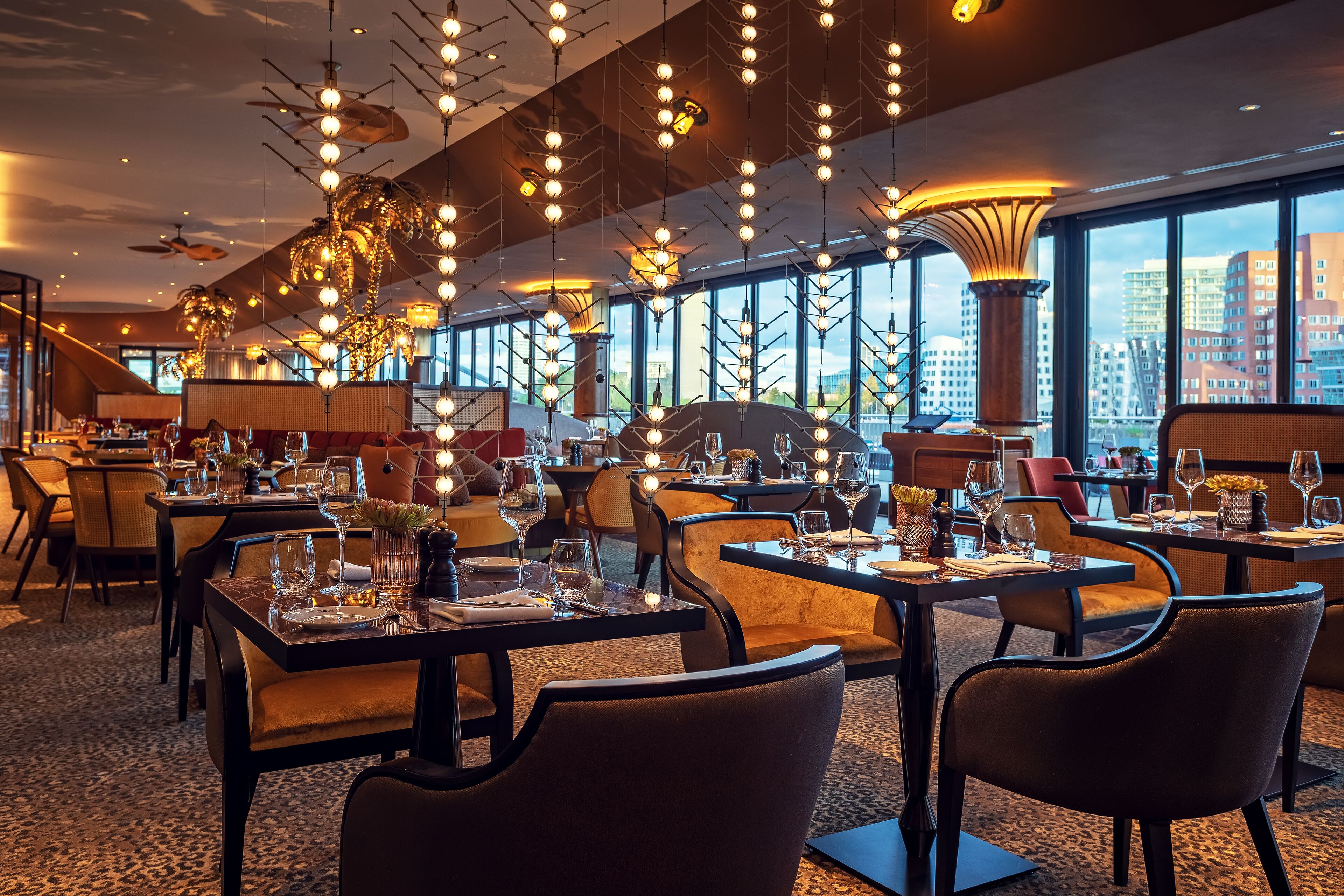 A modern restaurant with high ceilings, hanging spherical light fixtures, and large windows offering city views. Tables are set with wine glasses and cutlery.