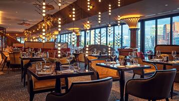A modern restaurant with high ceilings, hanging spherical light fixtures, and large windows offering city views. Tables are set with wine glasses and cutlery.