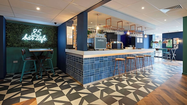 A bar area with a neon sign, bar stools, a counter with a drink dispenser, and a patterned floor. - Travelodge London Central Waterloo (London)