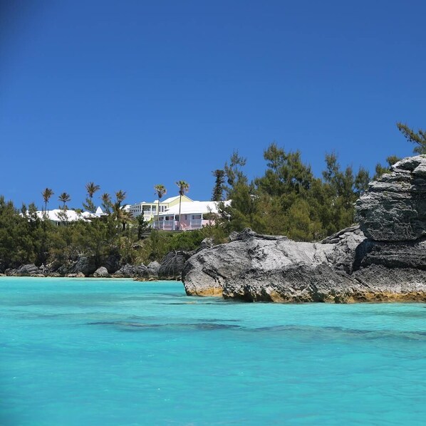 A coastal view with clear turquoise waters, a rocky outcrop, and a building with a white roof and multiple windows. - Willowbank Resort (Sandys Parish)
