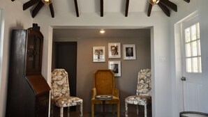 Una habitación con techo de madera, dos sillas estampadas, un sillón central y cuadros en la pared.
