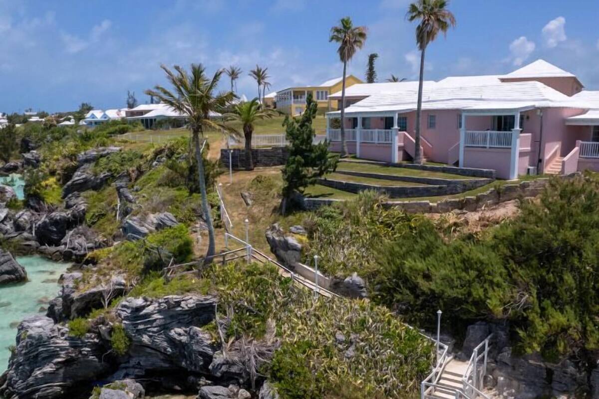 Uma propriedade à beira-mar com um edifício rosa, costa rochosa, água turquesa cristalina e palmeiras.
