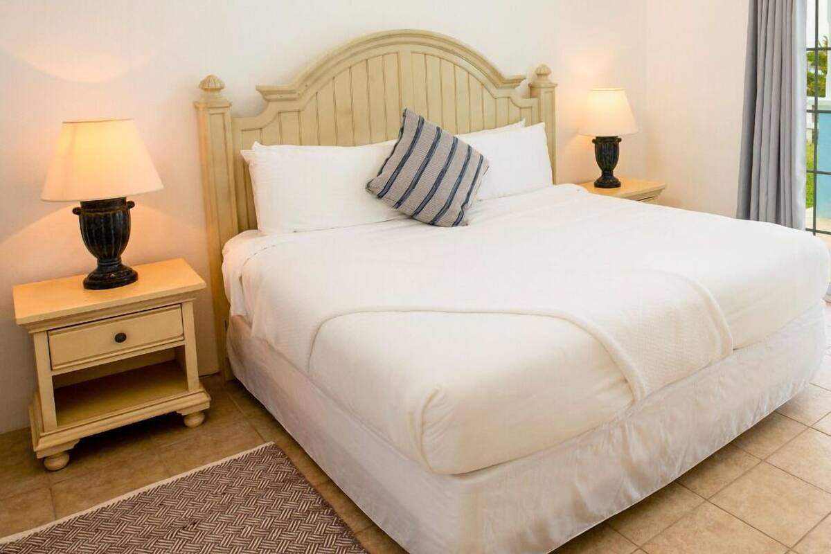 Uma cama bem arrumada com lençóis brancos e almofadas listradas, duas luminárias de cabeceira, uma pequena gaveta e um tapete com padrão herringbone.